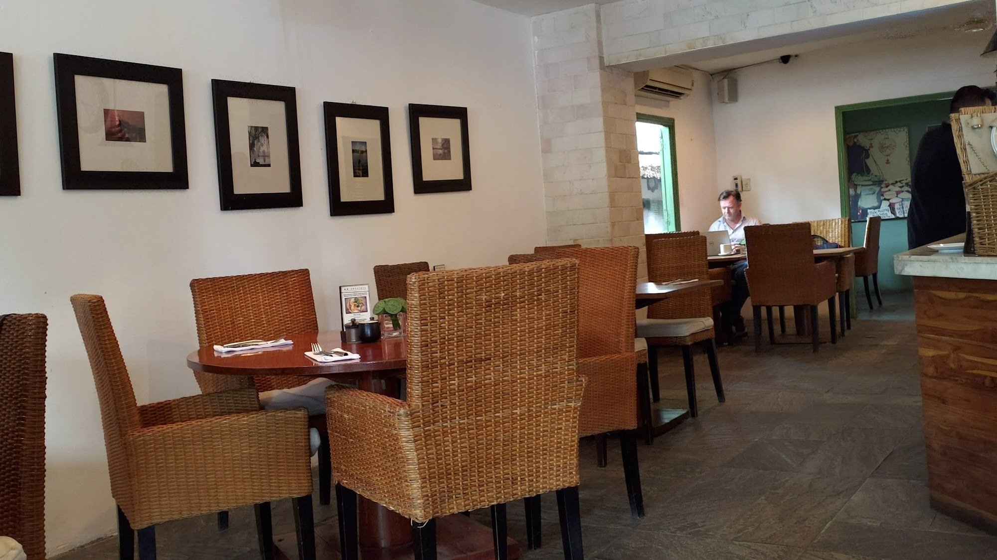 The image depicts the interior of a restaurant in Thao Dien, Saigon. It features wicker chairs arranged around wooden tables, framed black-and-white p