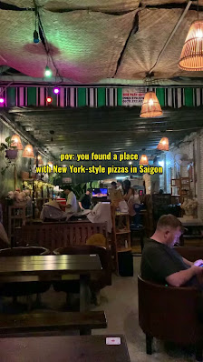 The image shows an indoor dining area in a restaurant or bar setting. Patrons are seated at tables with various activities, such as using their phones