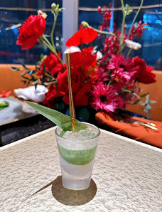 A close-up of a cocktail in a frosted glass with a green rim and garnish on top, placed against the backdrop of vibrant red flowers.