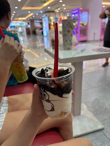 A person is holding a cup of dessert in an indoor setting that appears to be a shopping mall or food court with various shops and arcade games visible