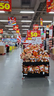 A well-lit supermarket interior in Thao Dien, Saigon, featuring a display of stuffed animals (teddy bears, koalas) on a black cart. Prominent red sign