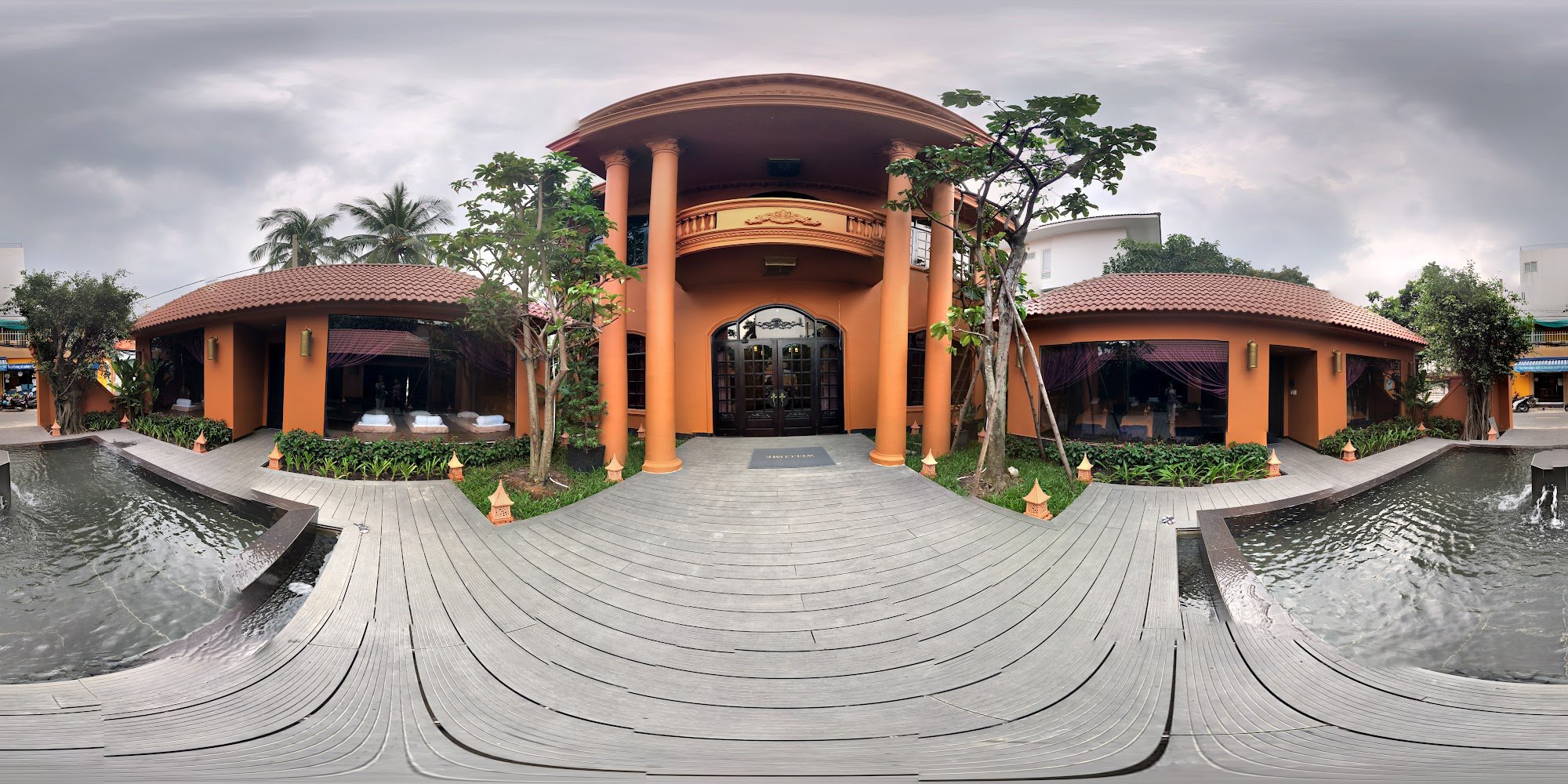 The image shows an exterior view of a luxurious building with terracotta-colored architecture and large pillars leading to the entrance, surrounded by