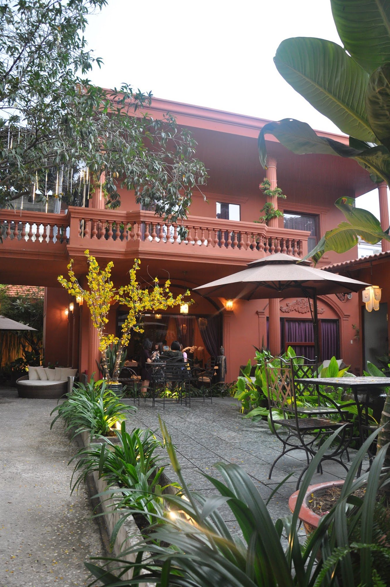 An exterior view of a two-story building with a reddish-brown facade, featuring a balcony with ornate railings, an outdoor seating area with tables, c