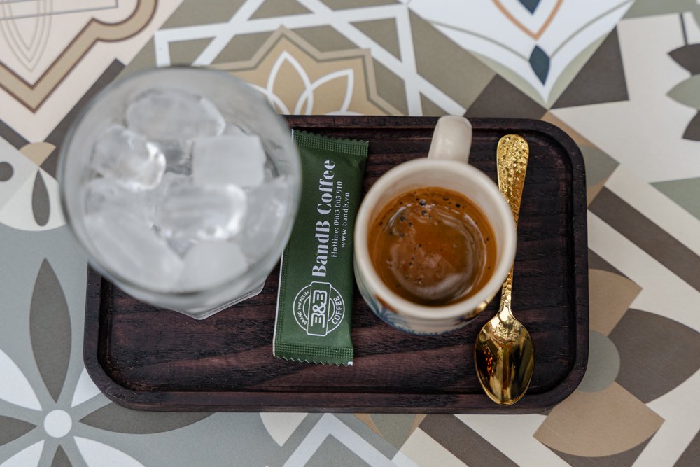 The image shows a close-up of a tray containing a cup of coffee and an empty glass with ice cubes on top, along with a packet of instant coffee mix an