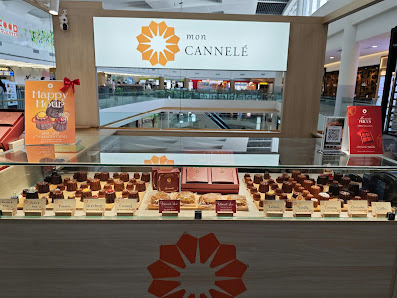 The image shows a display case in an interior setting of Thao Dien Mall featuring various chocolates and desserts from 'Mon Cannele'. The brand's logo