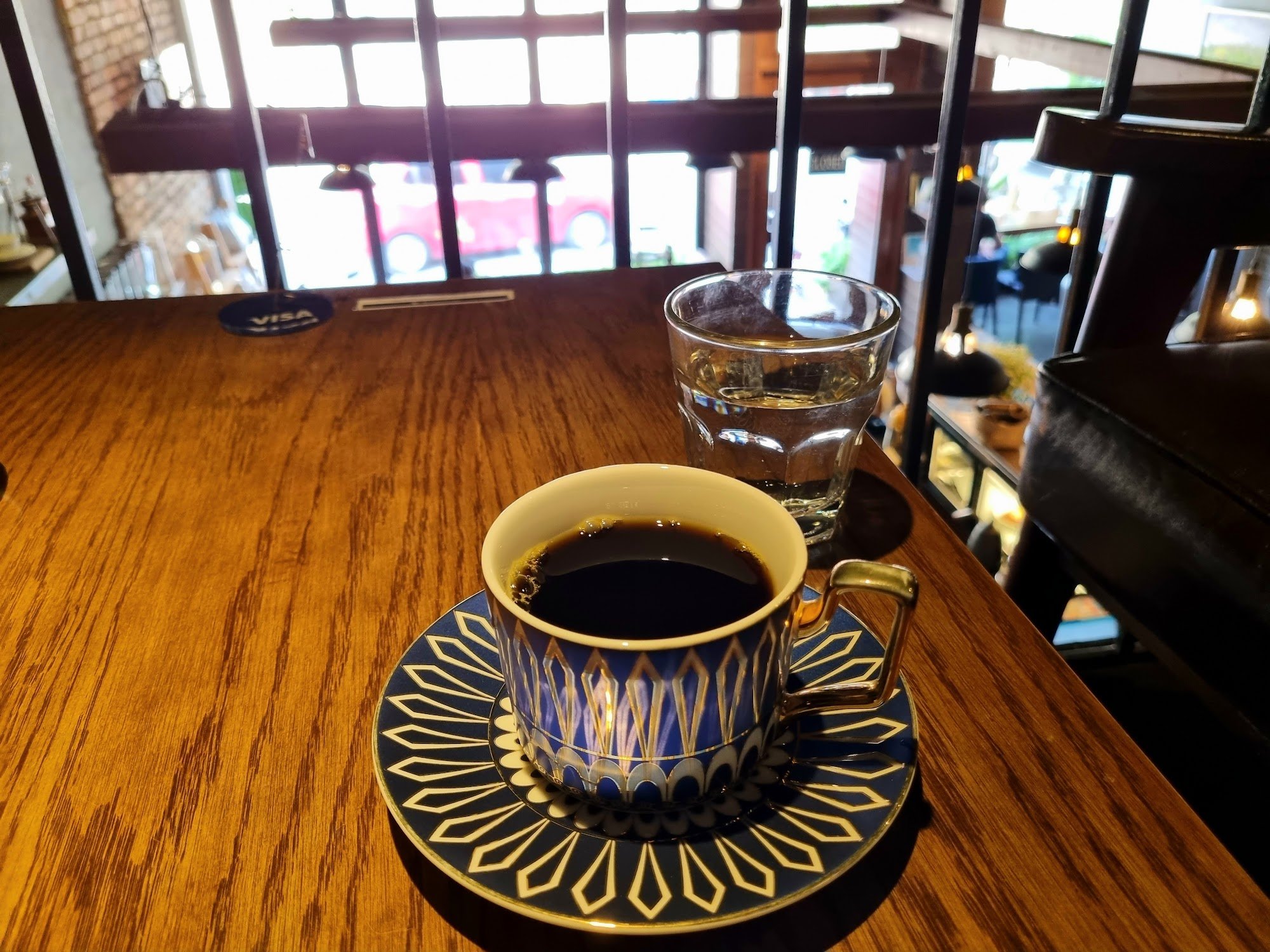 A close-up shot of a cup of coffee on an ornate saucer and plate with matching design, placed on a wooden table in what appears to be a cozy café sett