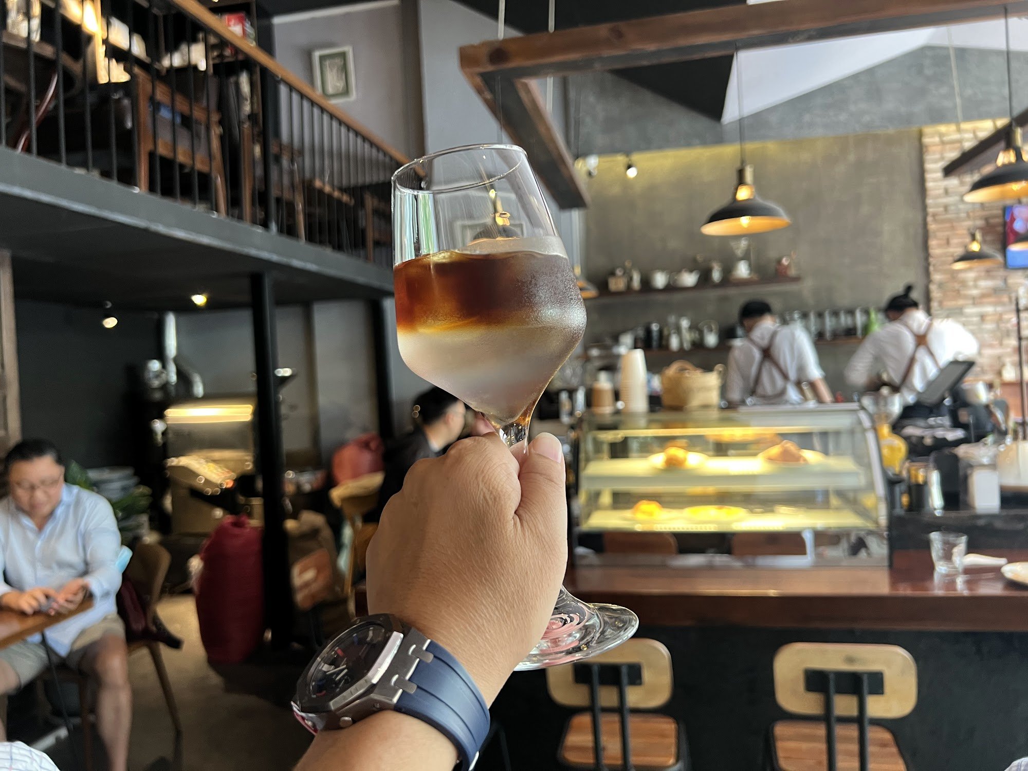 A person is holding a glass of coffee in an indoor setting that appears to be a café or restaurant with patrons seated and staff working behind the co