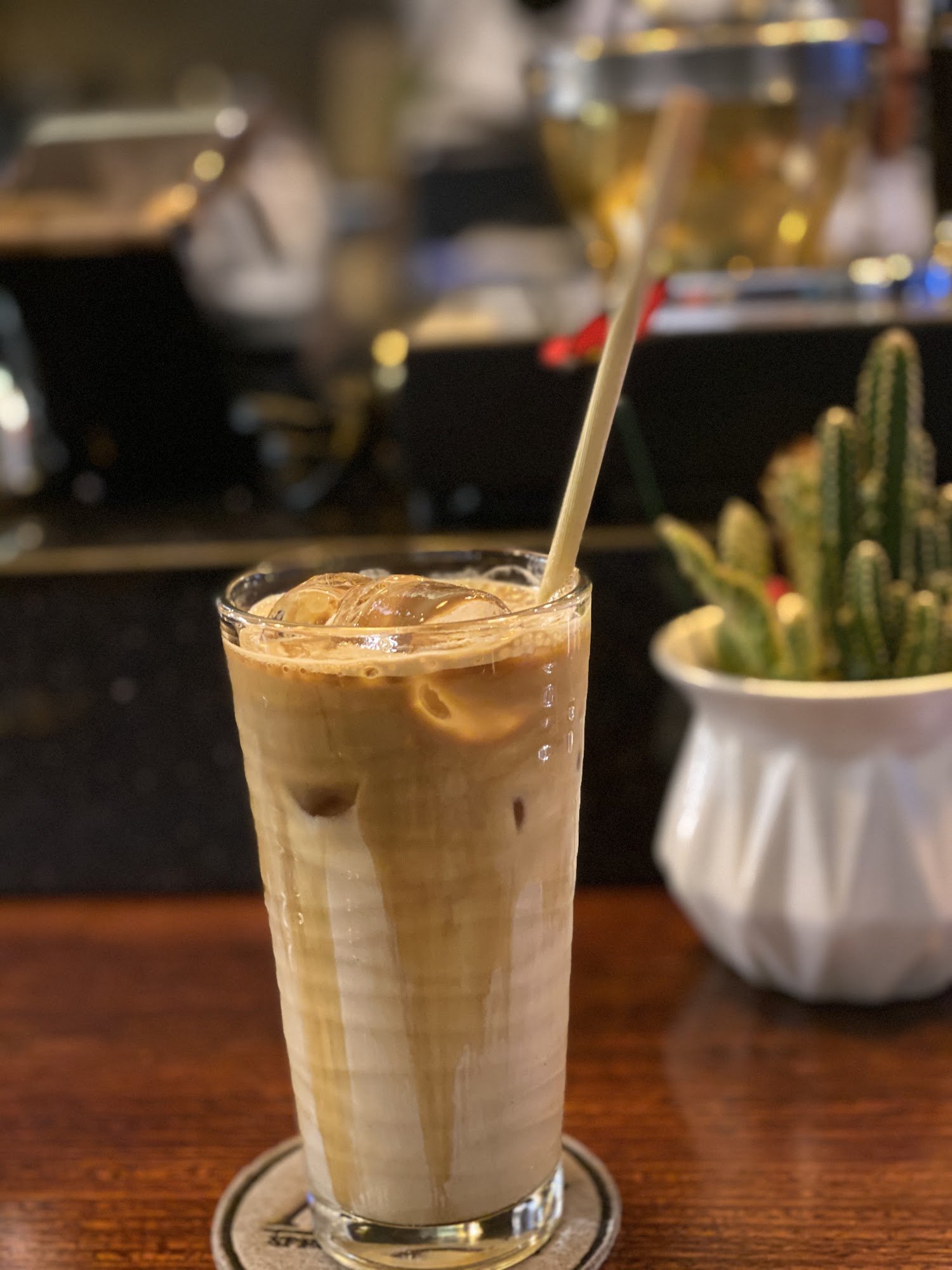 A glass of cold coffee beverage topped with a layer of milk foam and ice cubes on a wooden table in what appears to be an indoor setting, possibly a c