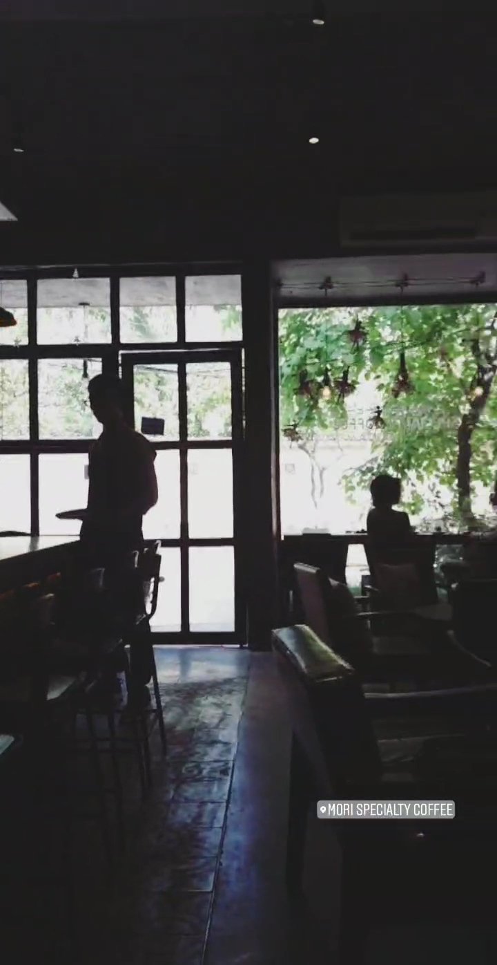 The image depicts an interior view of a coffee shop during the day. The lighting is dim with natural light coming through large windows, creating silh