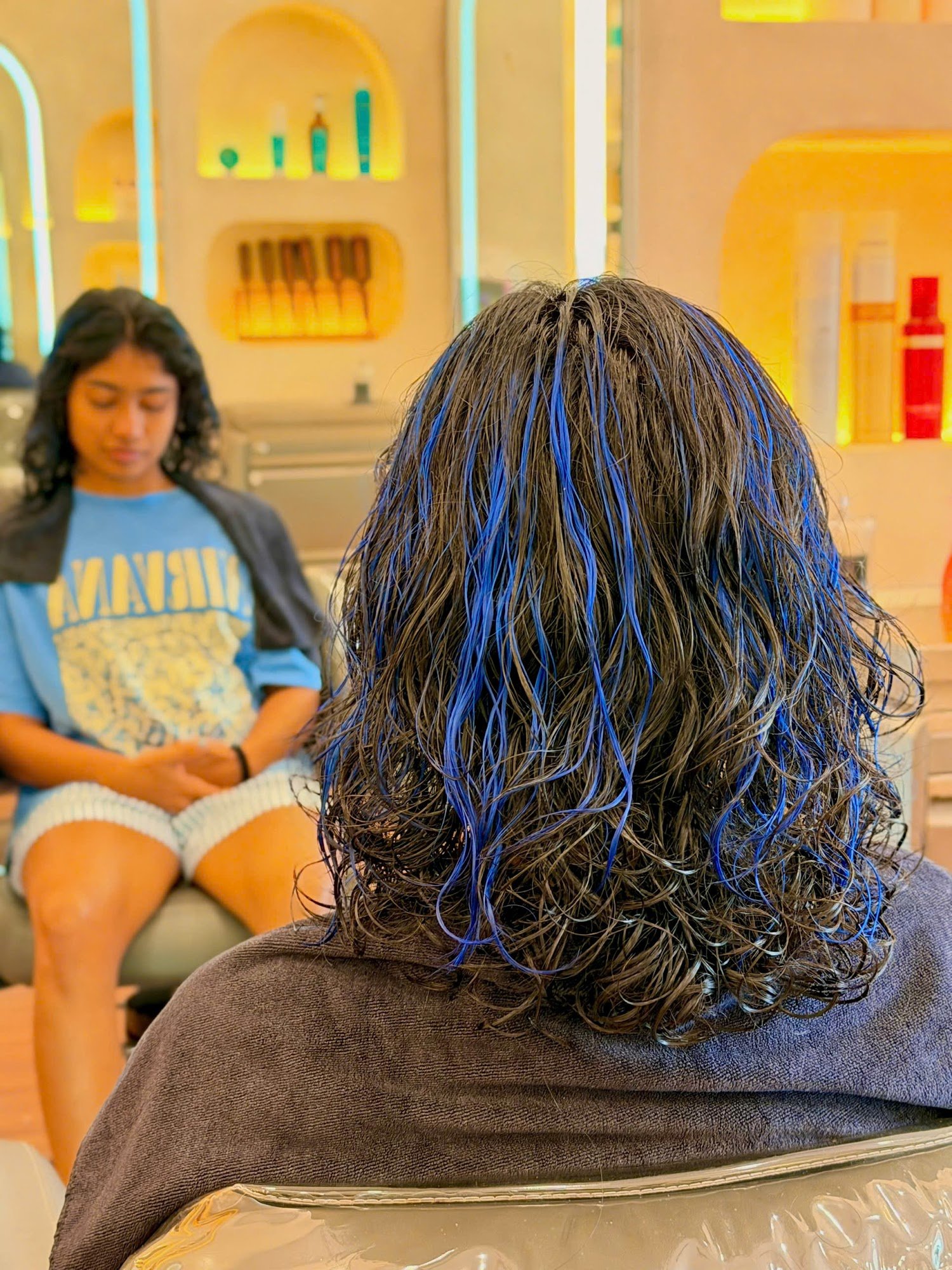 The image depicts an interior scene of a hair salon or beauty parlor where one individual is seated with their back to the camera, showing off blue st
