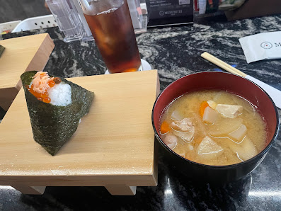 The image shows a meal consisting of sushi wrapped in seaweed and placed on a wooden block alongside a bowl of soup with visible ingredients such as v