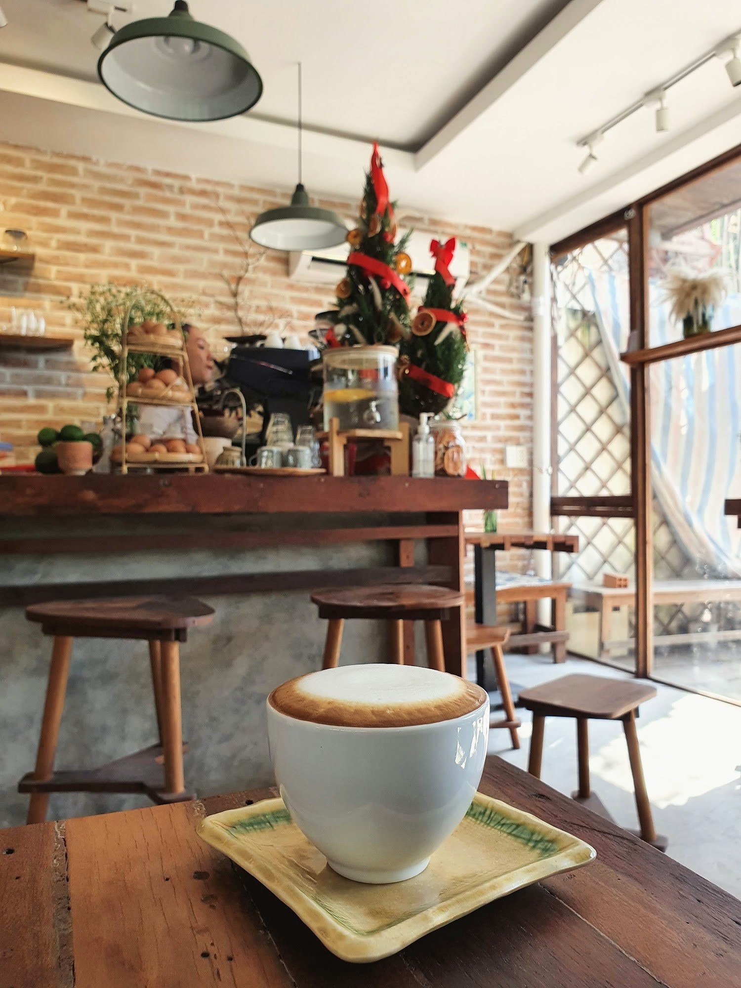 This image depicts an interior view of a cozy café or coffee shop during the holiday season. A cup of cappuccino is placed on a wooden table in the fo