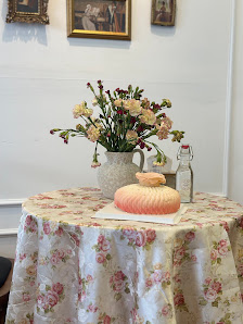 The image shows a table covered in a floral-patterned cloth on which sits a vase of flowers and a bottle with a label, alongside what appears to be a 