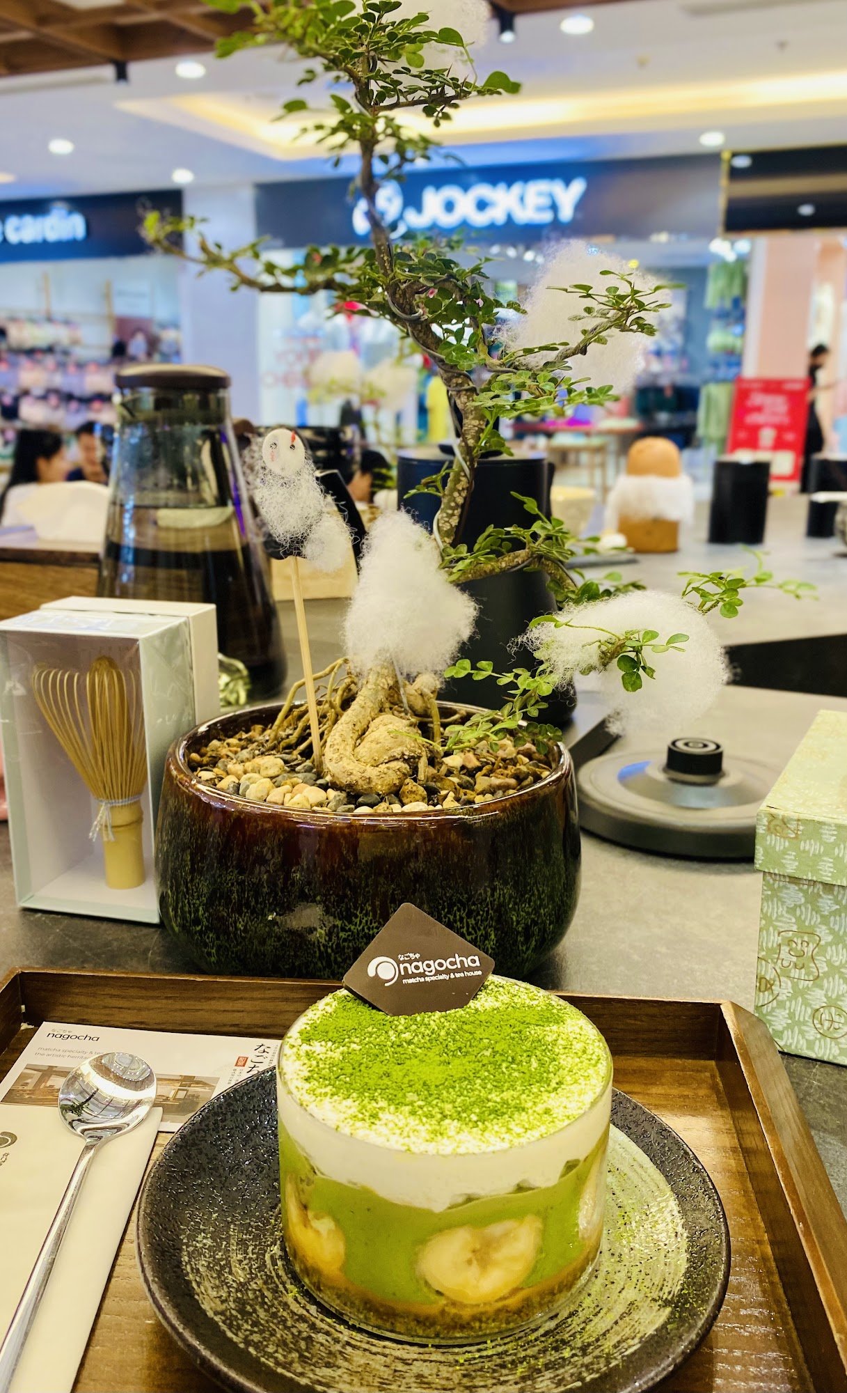 The image shows a dessert on a plate in the foreground, placed within an indoor setting that appears to be a café or restaurant with modern decor and
