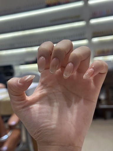 A close-up of a hand with neatly manicured, glossy, transparent nails, likely in a salon setting. Background shows blurred shelves, possibly in a stor