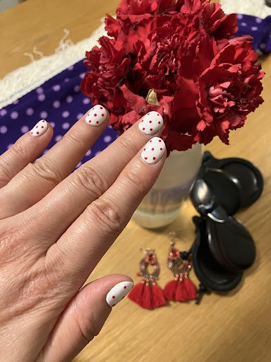 A close-up of a person's hand displaying manicured nails and accessories against the backdrop of an interior setting with red flowers in a vase, earri