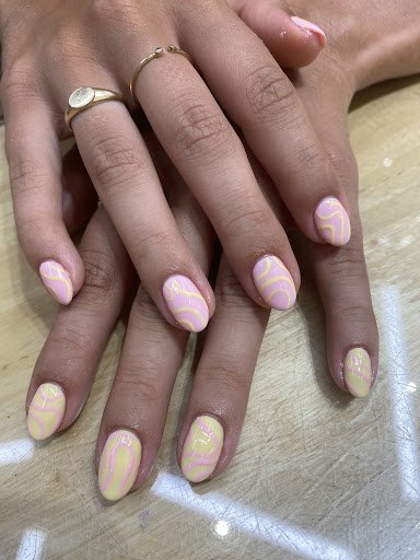 Close-up of two hands with manicured nails featuring a light pink and yellow swirl design. Nails are neatly shaped, and the person wears two gold ring