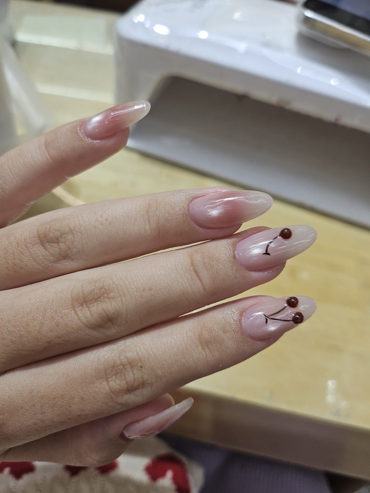 A close-up of a hand with long, almond-shaped nails featuring a glossy, nude-to-pink ombre design. Two nails have cherry blossom nail art with small b
