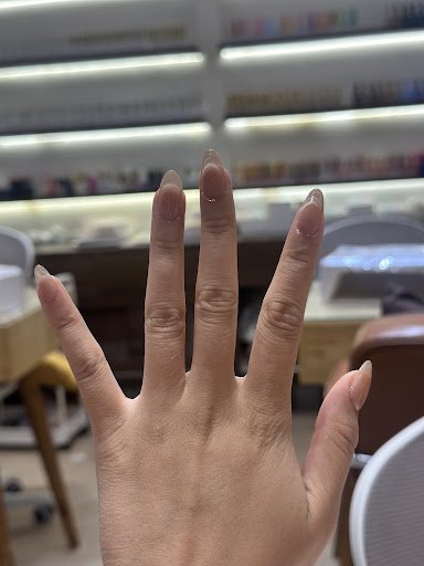 A close-up of a hand with neatly manicured nails, showing the fingers extended. The background features shelves with various products, likely beauty o