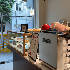 The image shows an interior view of a cafe or bakery shop featuring a display case with various pastries and desserts on the left, and a Rocket brande