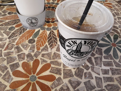 Two cups of beverages on a tiled surface; one is labeled 'New York Bagel & Coffee' with visible ice and straw, the other cup's label is partially obsc