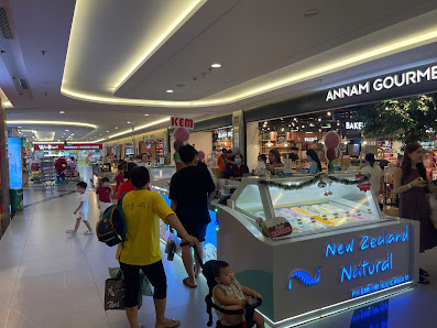 The image shows an interior view of a shopping mall or food court area featuring various stores and displays with people walking around.
