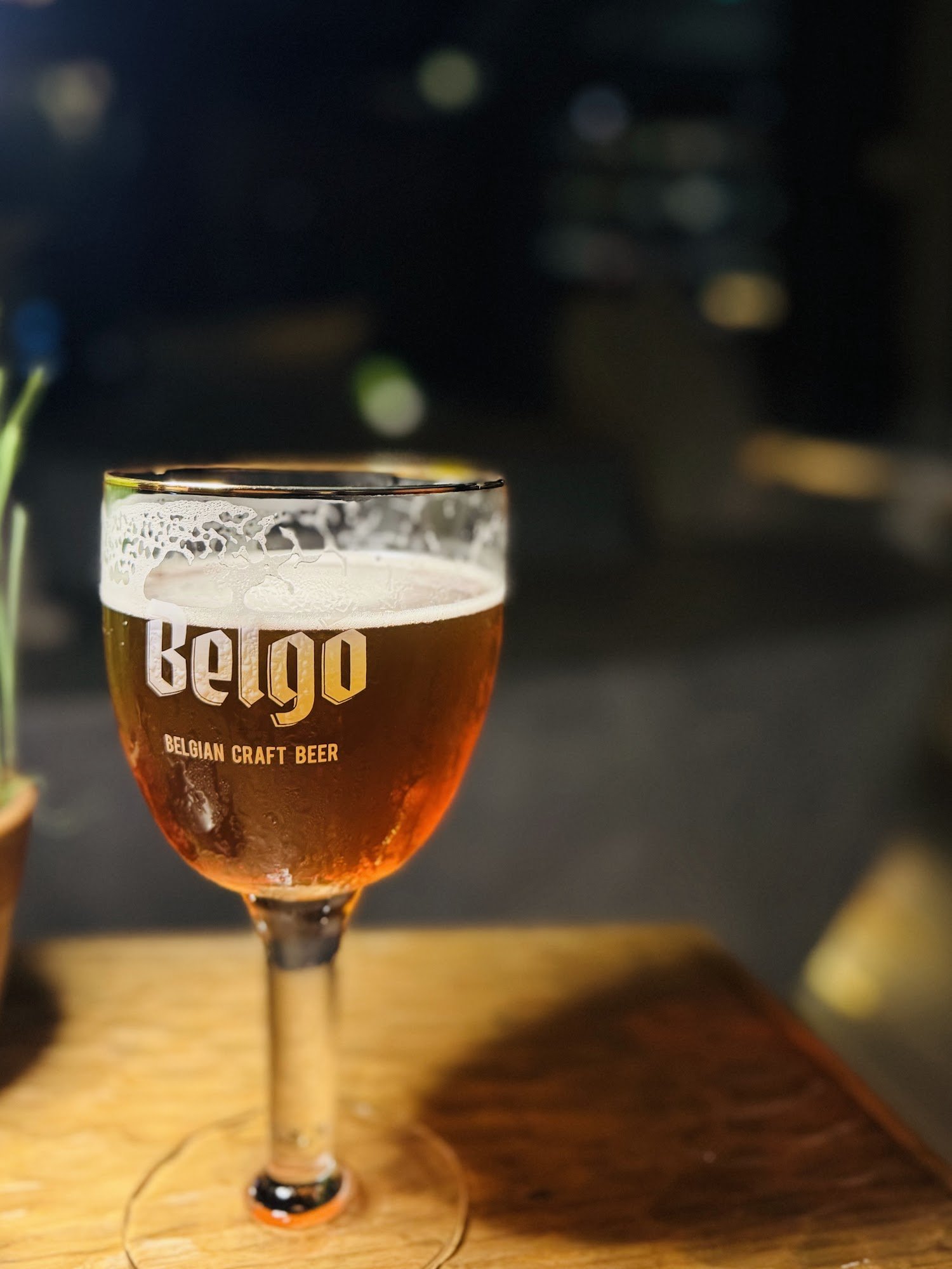 A close-up shot of a glass filled with amber-colored beer labeled 'Belgo Belgian Craft Beer'. The background is blurred, suggesting the photo was take