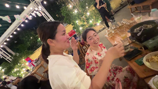Exterior view of a dining venue in Thao Dien, Saigon. Features outdoor seating with wooden tables, string lights, and greenery. Two women are dining, 
