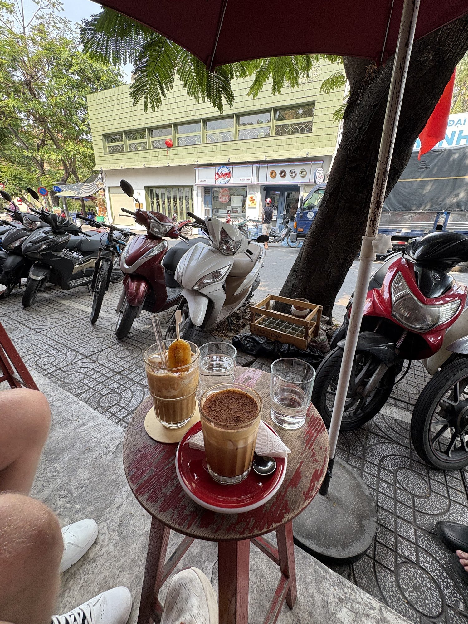 Outdoor café setting in Thao Dien, Saigon. A small round table with drinks: iced coffee with a corn dog, a cappuccino with cocoa powder, and two glass