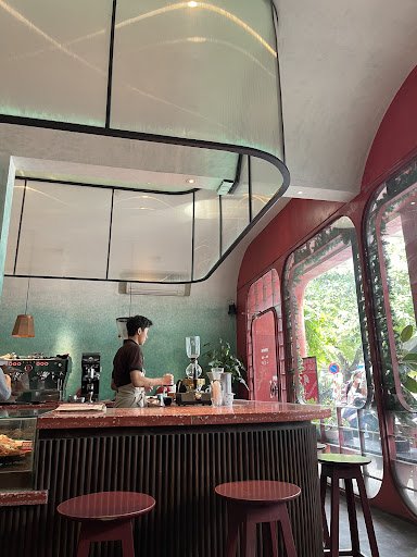 The image shows an interior view of a modern coffee shop or bar. A staff member is seen working behind the counter, which has a red and brown color sc