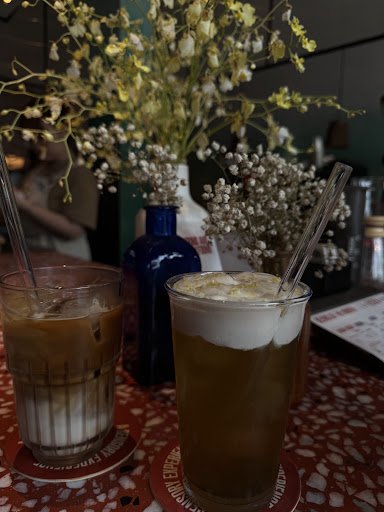 The image depicts a cozy interior setting of what appears to be a café or restaurant with two glasses containing cold beverages on the table, each gar