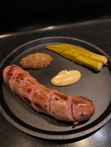 A plated dish featuring a sausage garnished with pink sauce and accompanied by mustard, pickles, and what appears to be fried chicken on the side.