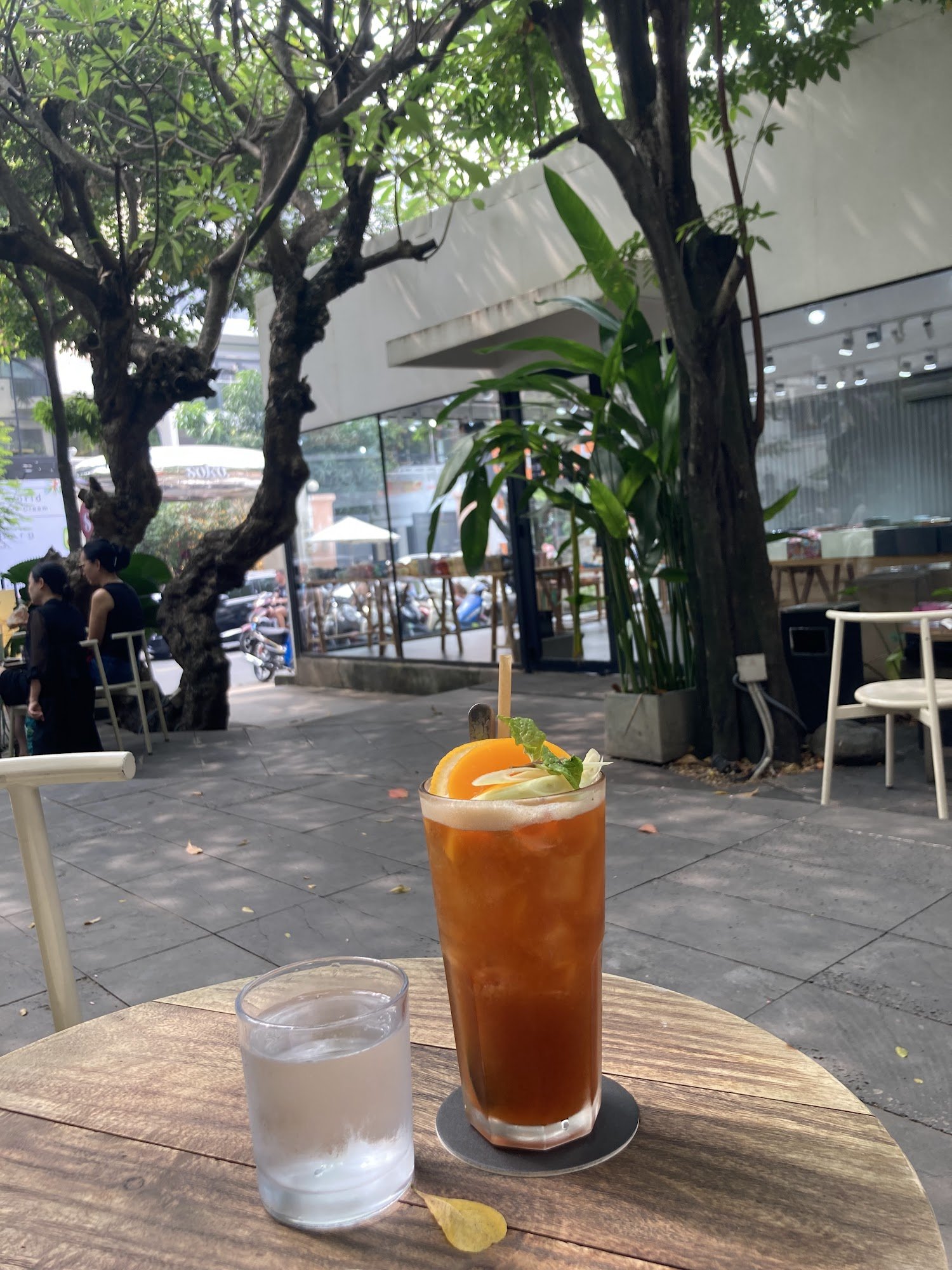 The image captures an outdoor seating area of a café or restaurant in Thao Dien, Saigon. In the foreground is a wooden table with two drinks on it: on