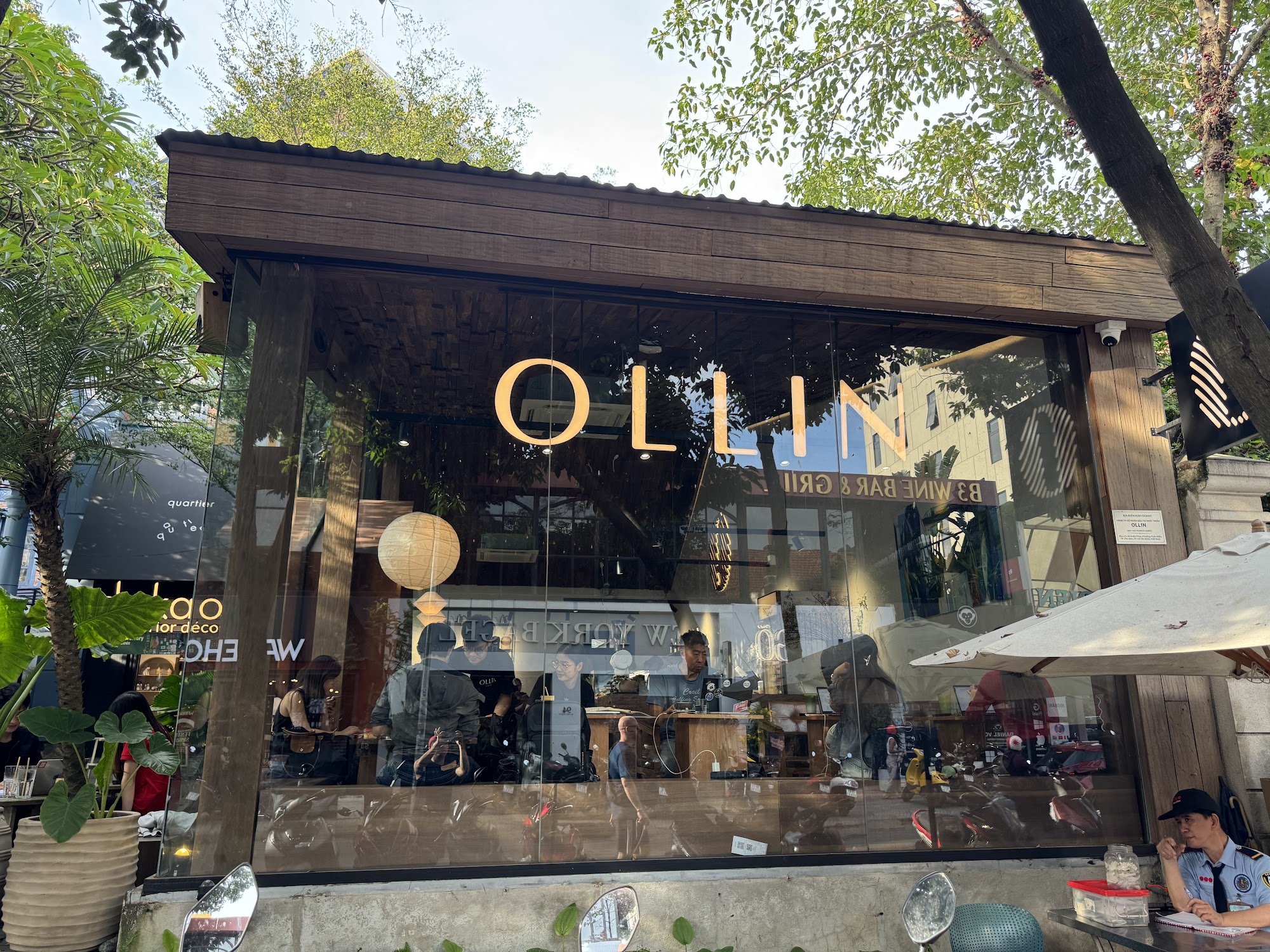 The image shows an exterior view of a cafe named 'OLLIN' located in Thao Dien, Saigon. The establishment has large glass windows displaying the name a