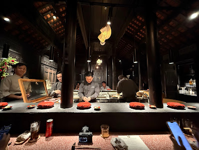 The image depicts an interior view of a restaurant or bar setting where patrons are seated at a counter with various dishes and drinks in front of the