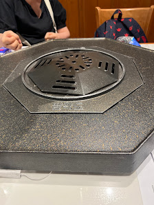 A close-up of a hexagonal, dark-colored device with a circular vent and 'HOT' branding, placed on a table. In the background, there are people (one we