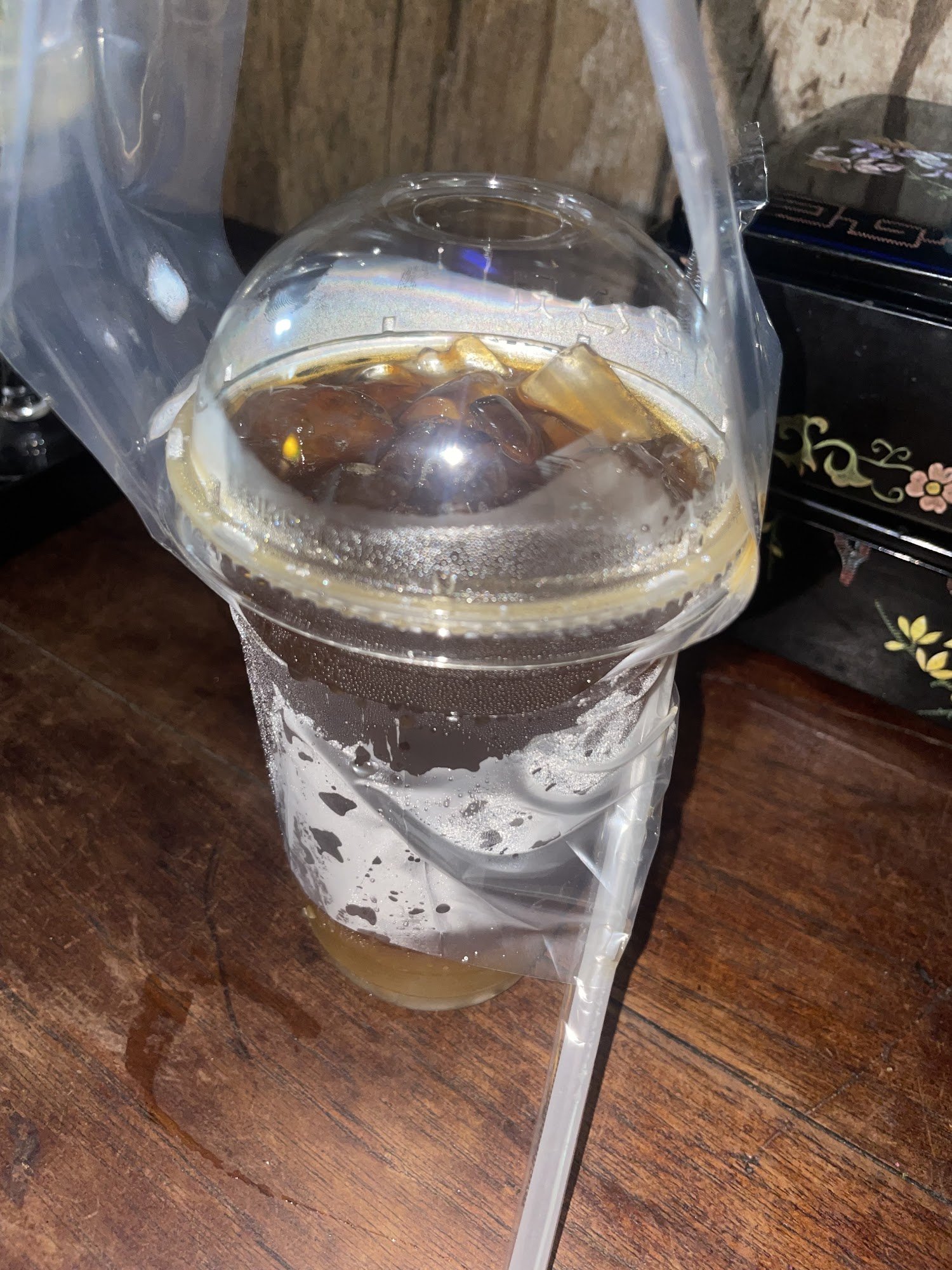 A close-up of a plastic cup containing an iced beverage with ice cubes and what appears to be syrup or condensed milk on the bottom, wrapped in clear 