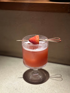 A close-up shot of a cocktail glass containing a pinkish-orange liquid garnished with a strawberry and two skewered straws.