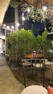 Outdoor seating area with round tables, metal chairs, potted plants, string lights, and a building with a concrete floor. A Coca-Cola bottle is placed