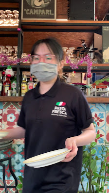 A staff member wearing a black polo shirt with 'ITALIA BECCA' logo, holding a plate, in a venue with shelves displaying various bottles, decorative it