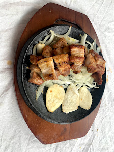 A dish consisting of pieces of pork belly and onions served on a black cast iron skillet placed on a wooden serving board.