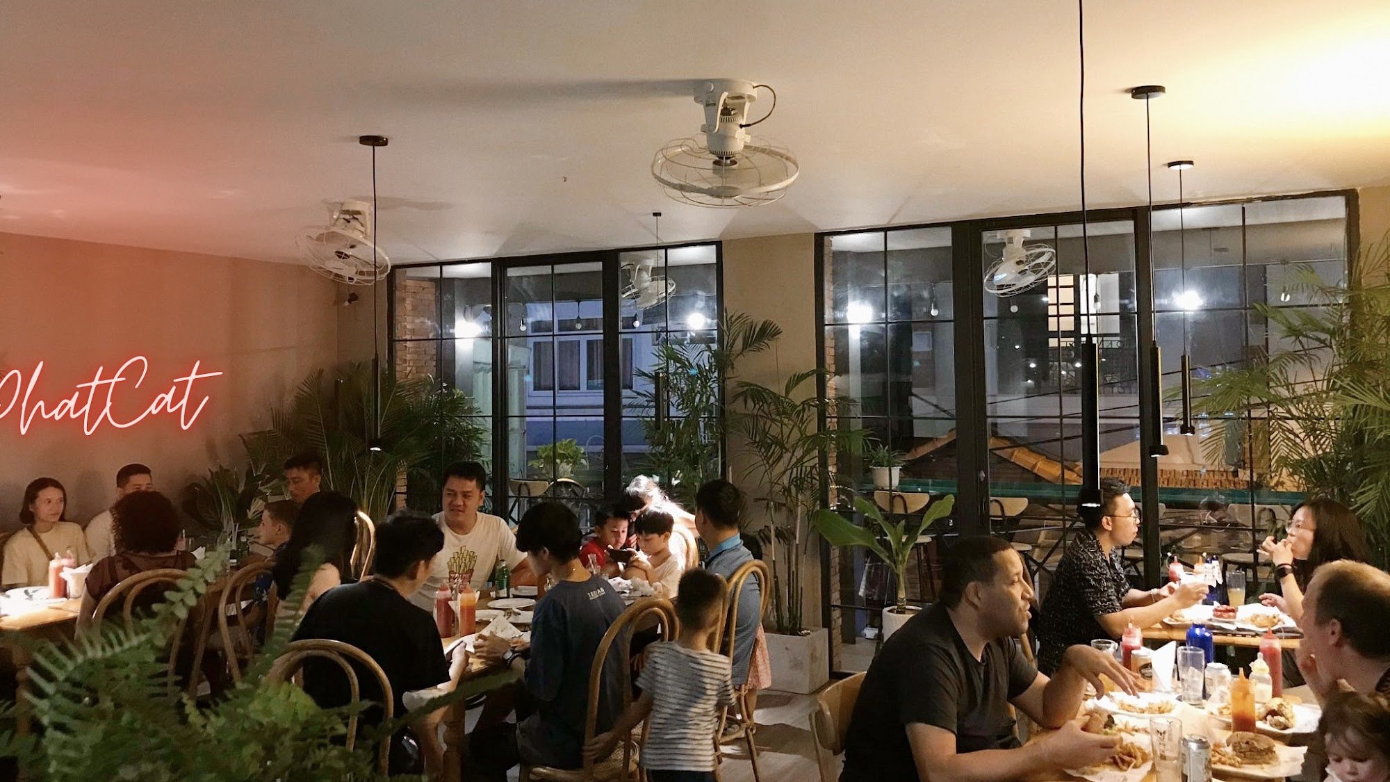 A bustling restaurant interior in Thao Dien, Saigon, with numerous patrons seated at tables. Features include large glass windows, hanging fans, potte