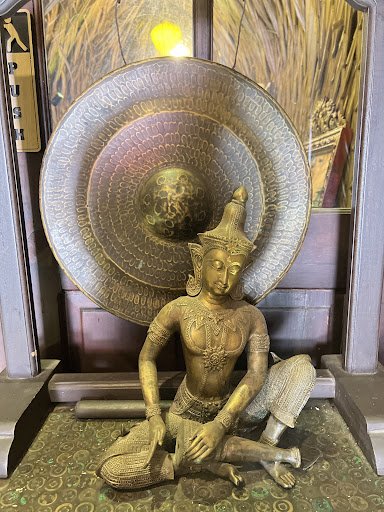 The image shows a brass statue of what appears to be an ancient figure seated in meditation pose against the backdrop of ornate, metallic decor includ