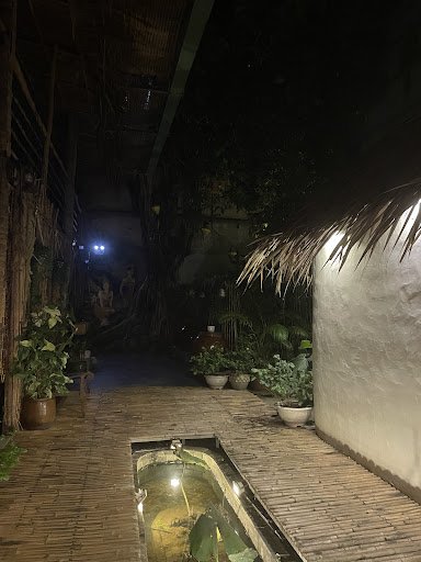 The image depicts an outdoor area of a venue at night time in Thao Dien, Saigon. It features a narrow pathway with bamboo flooring leading towards the