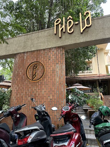 The image depicts the exterior of a venue named 'PHÊ-LA' in Thao Dien, Saigon. A concrete archway with a brick pillar features the venue's logo (a cir
