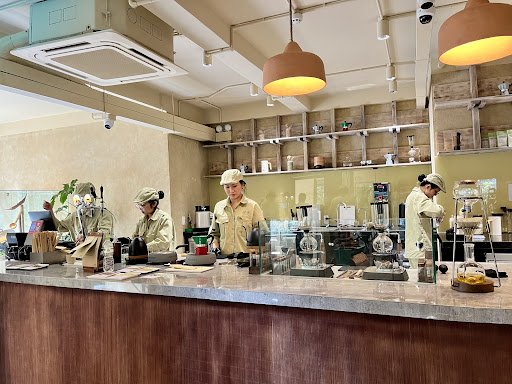 The image depicts the interior of a café or coffee shop in Thao Dien, Saigon. It features a counter with multiple staff members wearing uniforms and c