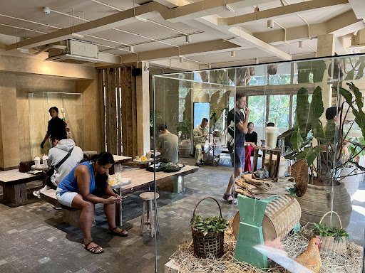 The image depicts an indoor venue in Thao Dien, Saigon, featuring a modern interior with wooden and glass elements. There are several people: a woman