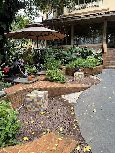 An outdoor patio area of a venue in Thao Dien, Saigon, featuring brick pathways, a large beige umbrella providing shade, potted plants and lush greene