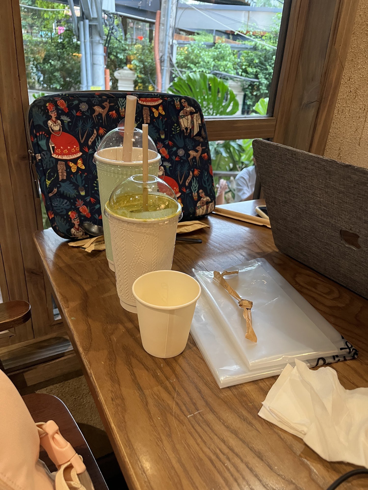 A wooden table in a café setting with two large cups with straws (one greenish, one yellowish), a small white cup, a plastic bag with a wrapped item,