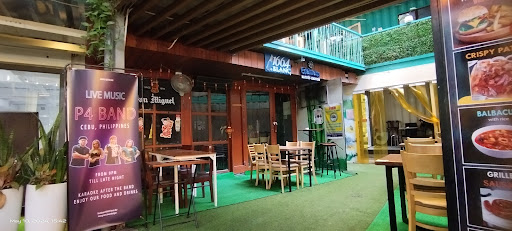 The image shows an outdoor seating area of a restaurant or bar named 'P4 Band'. It features wooden tables and chairs, green flooring, and various food